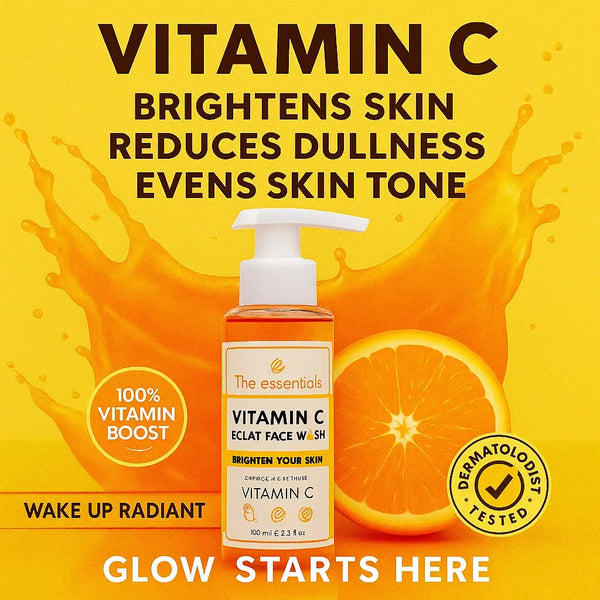 The Essentials Vitamin C Éclat Face Wash in a white pump bottle with orange-tinted gel, placed on a vibrant orange-yellow background with citrus splash, orange slice, and bold claims: brightens skin, reduces dullness, evens skin tone – labeled as dermatologist tested and 100% vitamin boost.